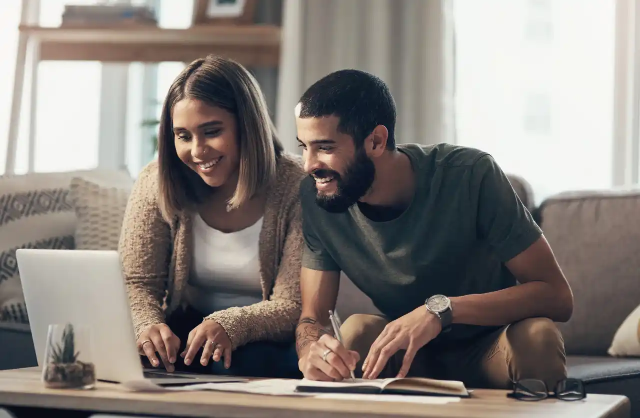 Casal sorridente fazendo planejamento de aposentadoria juntos em casa, usando notebook e anotando metas financeiras
