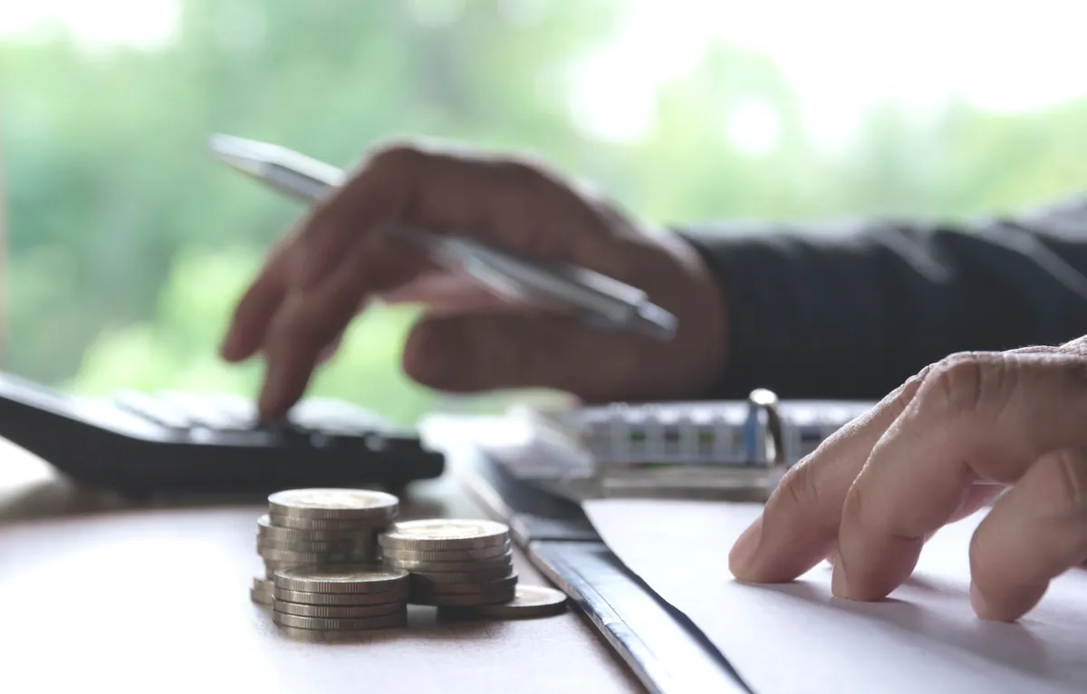 Mãos usando calculadora e caneta sobre pilhas de moedas em mesa de escritório