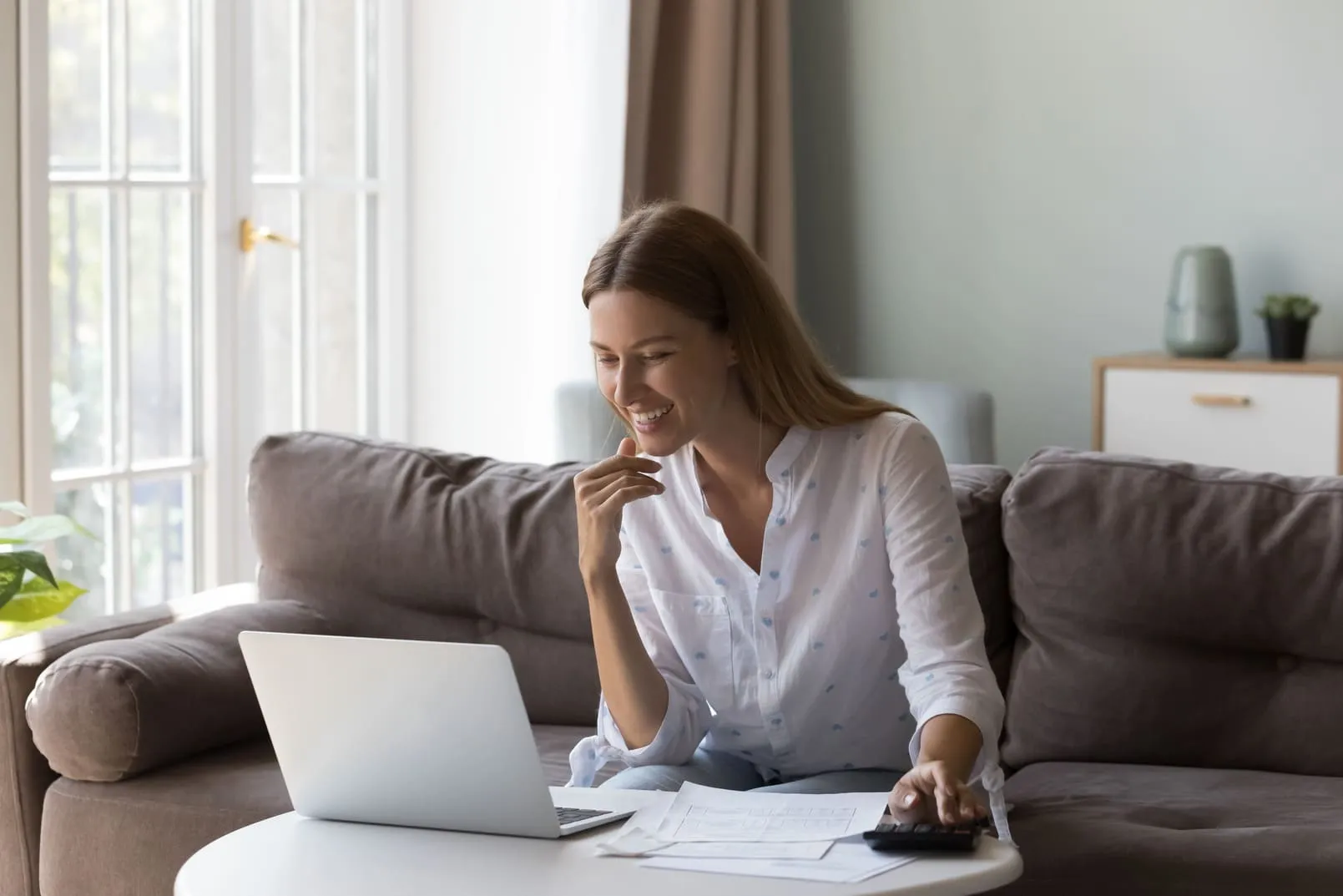 Mulher organiza orçamento pessoal no laptop em casa com planilhas e calculadora para controle financeiro.