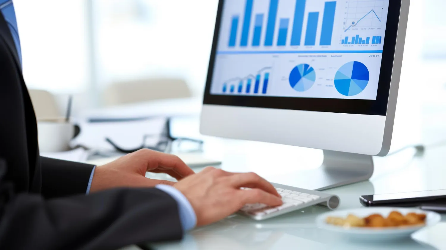 Pessoa digitando em teclado frente a monitor com gráficos e indicadores financeiros e análises visíveis.