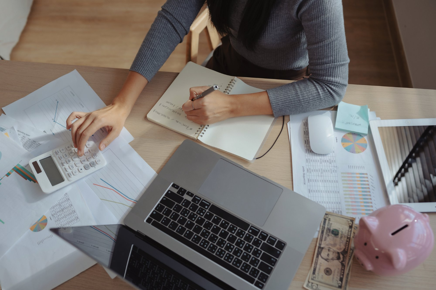 Como fazer um planejamento financeiro pessoal? Pessoa fazendo planejamento financeiro pessoal em um caderno, com calculadora, notebook, gráficos impressos e um cofrinho rosa sobre a mesa.