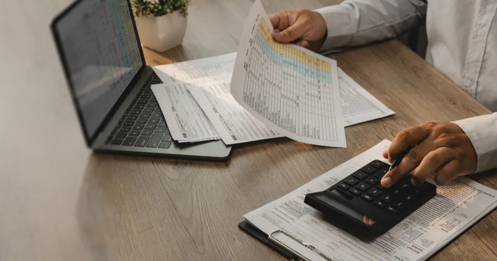 Mãos segurando documentos fiscais coloridos e calculadora sobre a mesa, ao lado de um notebook aberto, durante análise de regime tributário