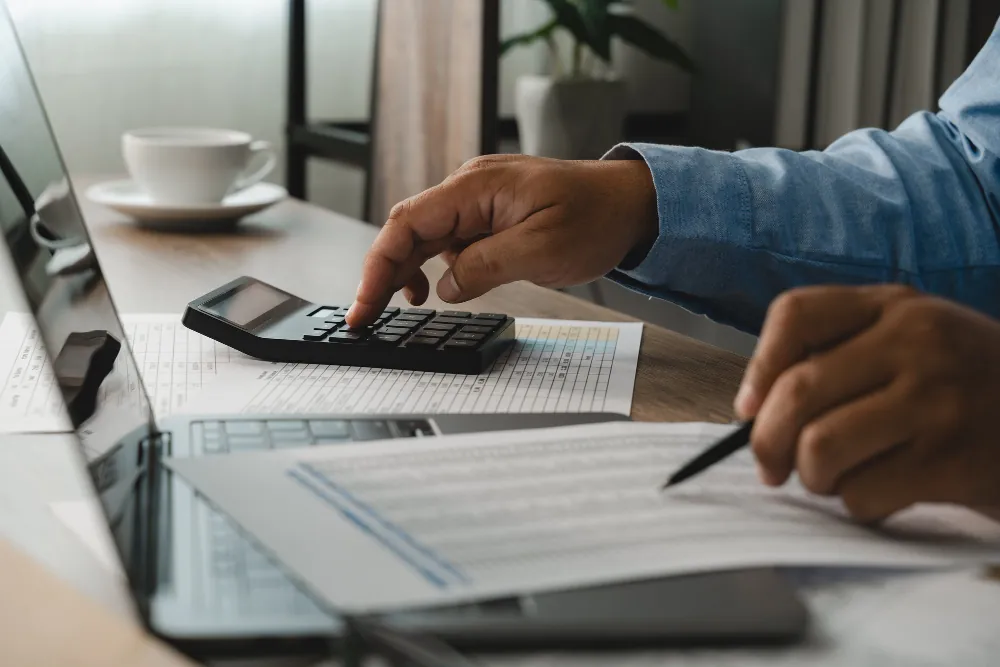 Mão usando calculadora sobre planilha de dados financeiros, ao lado de laptop e xícara de café, para cálculos de regime tributário.