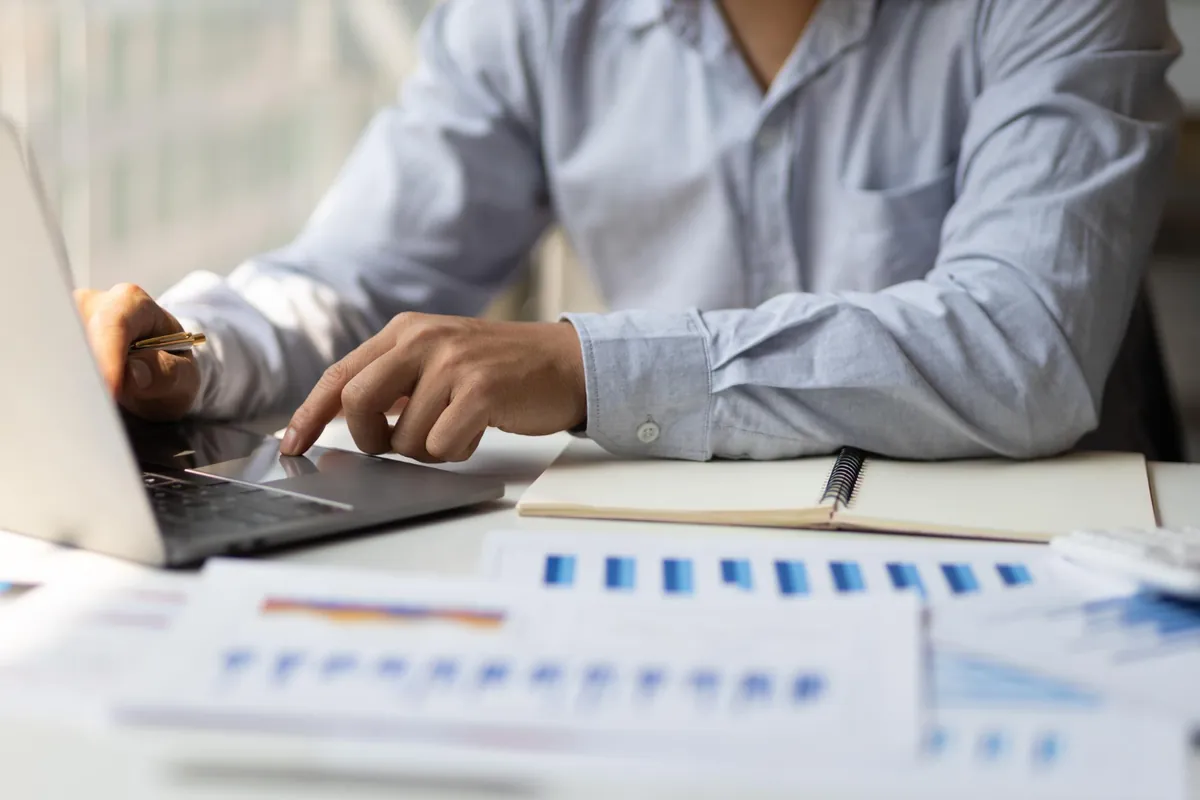 Profissional digitando no notebook ao lado de relatórios com gráficos financeiros impressos, atividade típica de um BPO financeiro