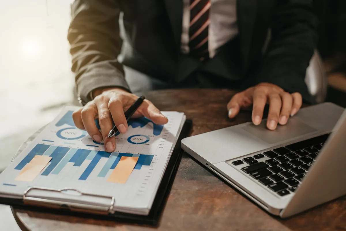 Como fazer diagnóstico financeiro empresarial? Executivo de terno apontando caneta para gráficos financeiros impressos ao lado de notebook, realizando diagnóstico financeiro empresarial