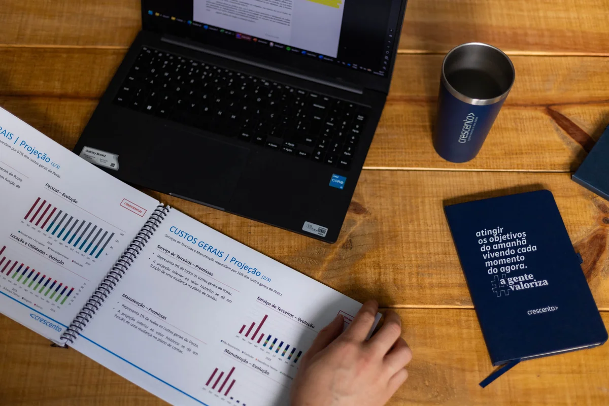 Diagnóstico financeiro empresarial: entenda sua importância antes de contratar uma consultoria e como fazer Relatório de diagnóstico financeiro empresarial aberto com gráficos de custos gerais, ao lado de notebook, copo térmico e caderno